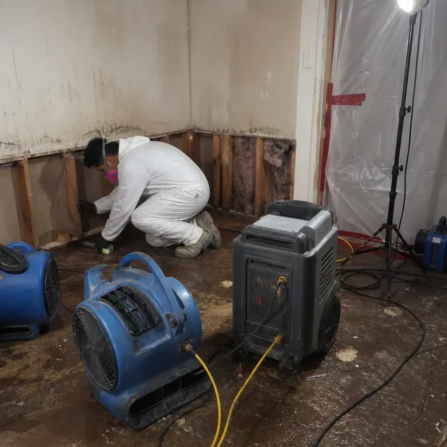 Tech in protective gear removing wet drywall beside air movers and a dehumidifier